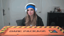 a woman wearing headphones sits in front of an orange box that says dare package