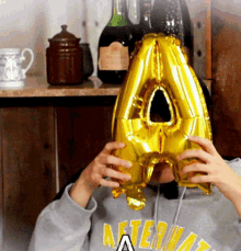 a person wearing a grey aftermath sweatshirt is holding a yellow letter a balloon in front of their face