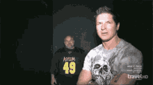 a man wearing an asylum 49 shirt stands in front of another man