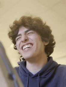 a young man with curly hair is laughing and wearing a blue hoodie