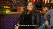 a woman in a leather jacket is sitting in front of a bookshelf and saying i bring the drama and the pizza .
