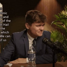 a man in a suit and tie sits in front of a microphone with a bottle of water next to him