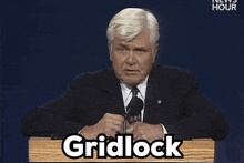 a man in a suit and tie stands at a podium with the word gridlock written on it