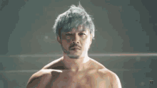 a shirtless man stands with his arms crossed in front of a blurred background with the letter t on it