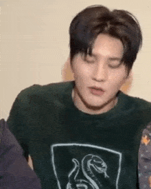 a young man wearing a green sweater with a snake on it is looking at the camera .