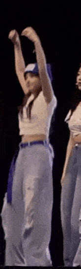two women are dancing on a stage with their arms in the air . one of the women is wearing a baseball cap .