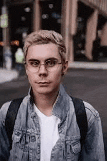 a young man wearing glasses and a denim jacket is walking down a street .