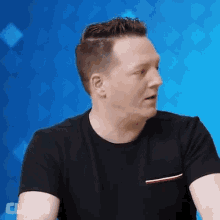 a man in a black t-shirt is sitting in front of a blue background with the word clash on it .