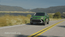a green car is driving down a road with mountains in the background