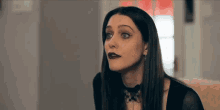 a woman wearing black lipstick and a choker is sitting on a couch and looking at the camera .