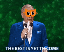a man in a blue suit and bow tie is holding a microphone and wearing a mask that says the best is yet to come