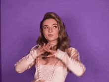 a woman in a pink dress is standing in front of a purple wall with her hands behind her head .