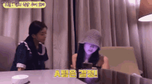 two girls are sitting at a table and one of them is wearing a purple hat .