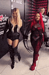 two women are walking down a hallway holding hands . one of the women is wearing a red wig .