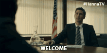 a man in a suit and tie sits at a table with the words welcome written on the table