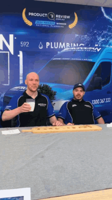 two men are sitting at a table in front of a plumbing van