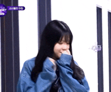 a girl with long black hair is wearing a blue jacket and covering her face with her hands .