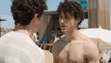 a shirtless man talking to another shirtless man on a beach