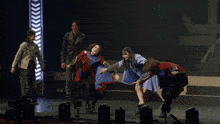 a group of people are dancing on a stage with a man in a black jacket standing behind them