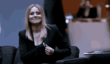 a woman in a black suit is sitting in a chair and smiling while giving a thumbs up .