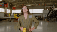 a man in a green jumpsuit stands in a hangar holding a yellow container that says ' honey ' on it