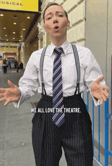 a man wearing suspenders and a tie stands in front of a building that says " we all love the theatre "