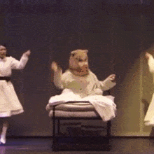 a teddy bear is sitting on a chair on a stage while two women dance behind him .