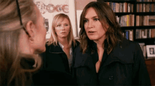 three women are standing next to each other in front of a bookshelf with a picture of a man on it