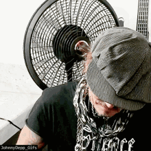 a man wearing a hat and a johnny depp gifs shirt stands in front of a fan