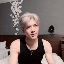 a man wearing a black tank top and a necklace sits on a bed
