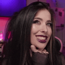a woman with long black hair and purple lipstick is smiling and holding her hand to her face .