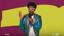 a man wearing a blue jacket and white glasses stands in front of a microphone with the nbc logo behind him