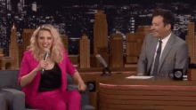 a woman in a pink suit is sitting in front of a microphone while a man in a suit sits behind her .
