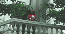 a man in a red jacket is standing on a balcony with trees in the background .