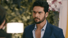 a man with a beard is wearing a suit and a blue shirt and looking at another man .