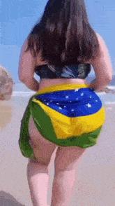 a woman in a bikini is walking on a beach with a brazilian flag draped over her hips .