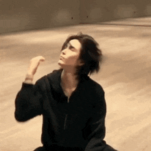 a man in a black hoodie is sitting on the floor in a room and making a funny face .