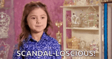a little girl in a blue shirt is standing in front of a shelf and says `` scandal-loscious '' .
