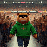 a teddy bear wearing a green hoodie and sunglasses stands in front of a crowd of people