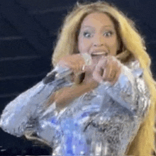 a woman is holding a microphone and pointing at the camera while wearing a silver dress .