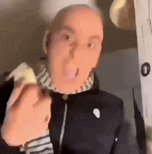 a bald man wearing a black jacket and a striped shirt is eating a banana .