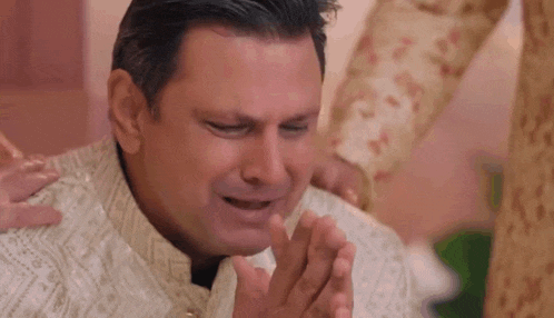 a man is crying while sitting on the floor with his hands folded in front of his face .