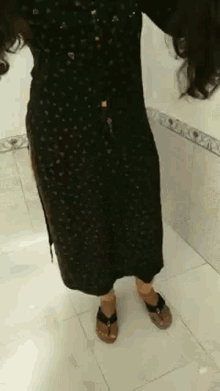a woman in a black dress and sandals is standing in a bathroom .