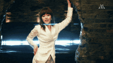 a woman in a white jacket stands in front of a brick wall with the letter m above her
