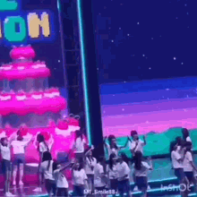 a group of young women are dancing on a stage in front of a large pink cake .