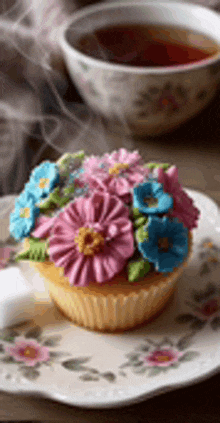 a cupcake decorated with pink and blue flowers on a plate next to a cup of tea .