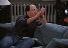 a bald man wearing glasses is sitting on a couch with his hands in the air