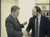two men in suits shake hands in front of a shelf with a plate on it