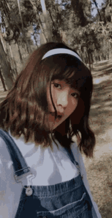 a girl wearing overalls and a white headband stands in a forest