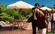 a man holds a woman in his arms in front of a table and chairs under an umbrella